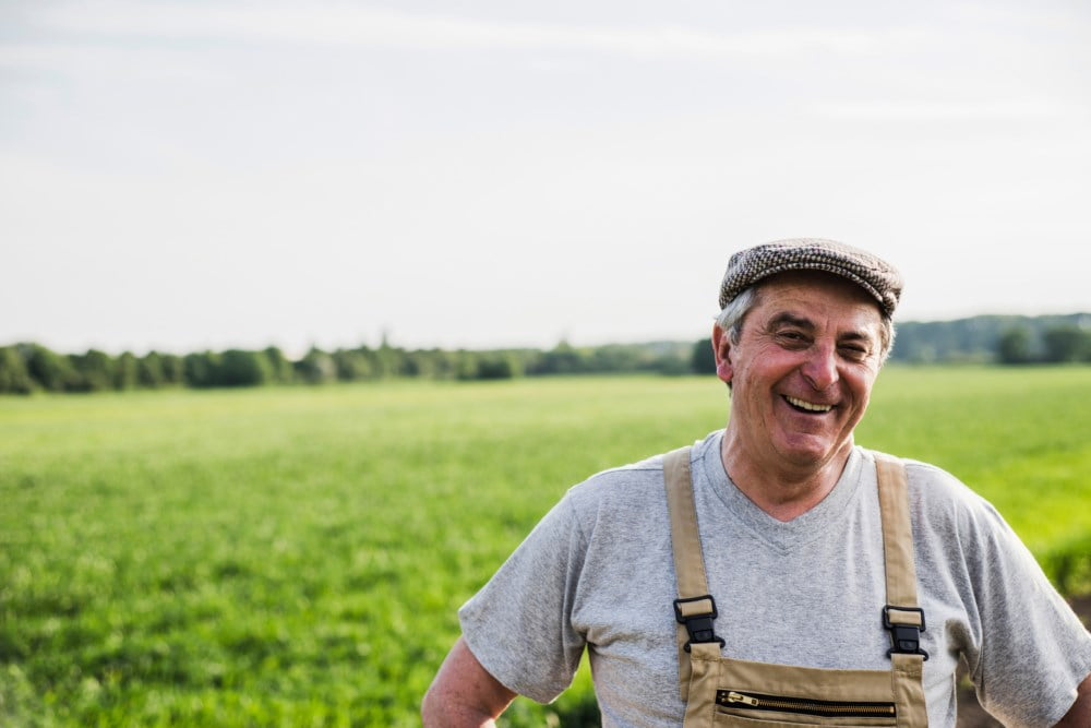 En eldre bonde med et bredt smil, kledd i hvitt t-skjorte og overalls, står i et stort, åpent grønt felt. Han har på seg en flat cap og ser glad ut mot kameraet på en solrik dag.