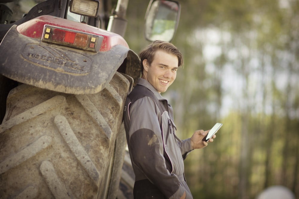 En ung bonde i en mørk kjeledress smiler og ser på kameraet mens han holder en smarttelefon, lent mot en stor landbruksmaskin, spesifikt en skitten rød traktor. Bildet er tatt utendørs i et skogkantet område, noe som gir en naturlig og arbeidsom bakgrunn.