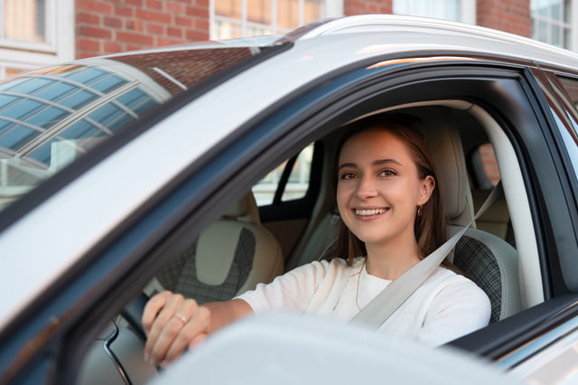 Ung kvinne smilende bak rattet i en firmabil, klar til å kjøre. Hun er sikkerhetsbeltet og ser ut gjennom frontruten, med en bakgrunn som antyder et boligområde. Dette bildet viser en behagelig og profesjonell bruk av en næringsbil.